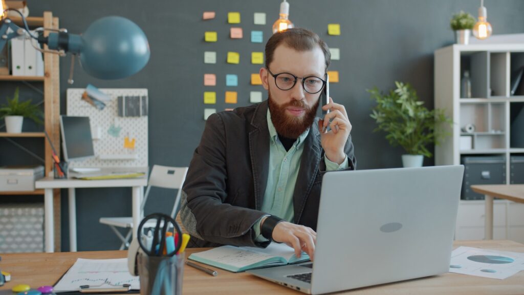 Dirigeant de PME au téléphone à son bureau, ordinateur portable ouvert et carnet de notes, scène d appel de prospection commerciale