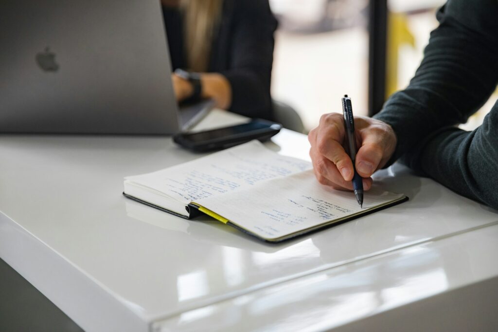 Carnet de notes ouvert sur un bureau avec un MacBook au fond, scène de debrief commercial après une mission de prospection B2B