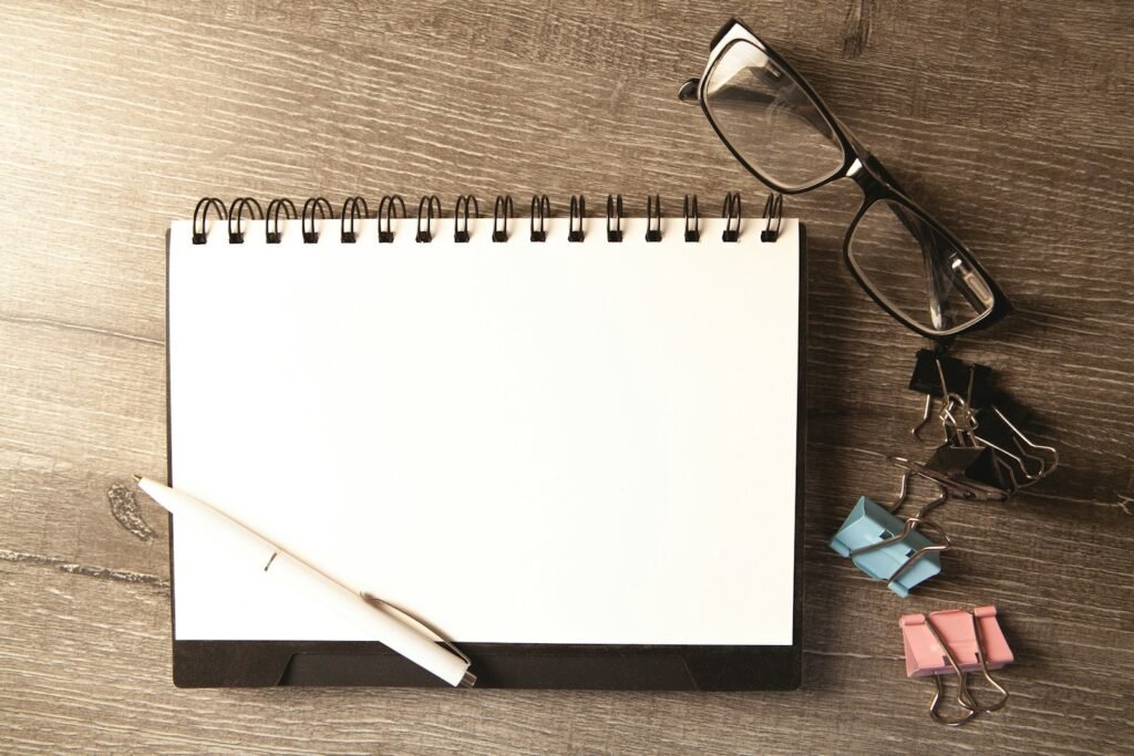 Un carnet à spirale ouvert sur une page blanche, un stylo et une paire de lunettes posés sur un bureau en bois